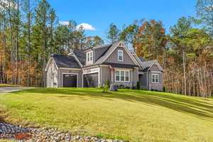 Single-story gray house with a gabled roof, attached garage, and stone accents, set on a sloped grassy lawn with trees in the background under a blue sky.