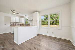 Bright kitchen and dining area with white walls, wood-style flooring, white cabinets, and a kitchen island. Large windows provide a view of trees and a grassy yard outside. Ceiling fans and modern lighting are visible.