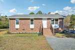 A single-story brick house in Oxford at 4158 Shock Overton Road features a gray shingle roof, white door, three front windows, brick steps, a gravel driveway, and a wheelchair-accessible ramp. The grassy yard is dotted with small plants.