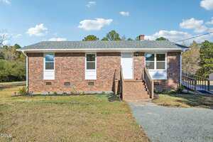 A single-story brick house in Oxford at 4158 Shock Overton Road features a gray shingle roof, white door, three front windows, brick steps, a gravel driveway, and a wheelchair-accessible ramp. The grassy yard is dotted with small plants.