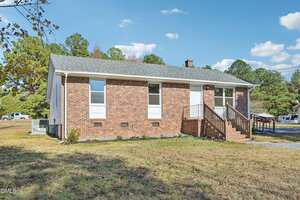 A single-story brick house with three tall windows and a white front door, accessed by a small set of stairs with a wooden railing. The yard has grass and scattered fallen leaves, with trees and a driveway in the background.