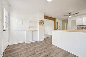Bright kitchen area with white cabinets, wood countertops, light wood flooring, white walls, open shelving, a ceiling fan, and a doorway leading to another room.