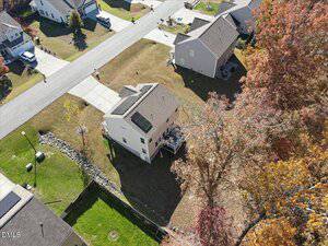 Aerial view of a suburban house with solar panels on the roof, a backyard deck, and neat lawn. Surrounding homes, trees with autumn foliage, and a road are visible.