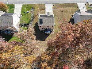 Aerial view of a suburban house with solar panels on the roof, a small backyard, and a deck. The house is bordered by lawns and trees with autumn foliage. A road and neighboring houses are visible nearby.