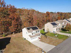 A two-story house with solar panels on the roof sits on a lawn next to a forested area. Nearby, other similar houses line the street under clear skies in a suburban neighborhood during autumn.
