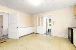 A spacious, empty kitchen with cream walls, a tiled ceiling, linoleum floors, white cabinets along one wall, an open door leading to another room, and a glass door to a sunroom or porch.