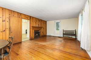 A living room with wood-paneled and cream-colored walls, hardwood floor, a fireplace, two windows with white curtains, a wooden bench, and part of a round dining table and chairs in the foreground.
