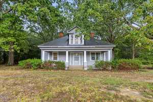 A small, light gray house at 1089 Gillburg Road in Henderson features a covered front porch, white trim, and a dormer window. It sits among tall trees and bushes with a patchy dirt and grass yard in front.
