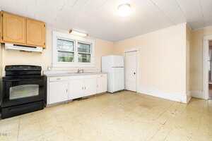 A simple kitchen with a black stove, white cabinets, a white refrigerator, and a small white door. There are two windows above the sink and beige walls with a tile floor.