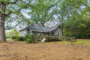 A gray, single-story house with a sloped roof, brick chimneys, and a wooden ramp leading to the porch. The house is surrounded by large trees, sparse grass, and a mostly bare dirt yard.
