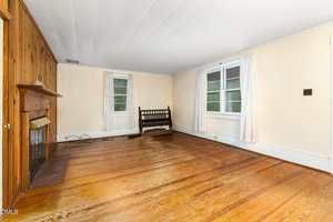 A mostly empty living room with wood floors, a wooden fireplace on the left wall, two windows with white curtains, cream-colored walls, and a wooden bench along the back wall.