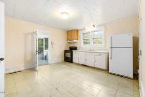 An empty kitchen with a white ceiling, light yellow walls, and a linoleum floor. The room has a stove, white refrigerator, cabinets, a window over the sink, and a door leading to an adjacent sunroom.