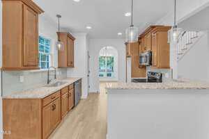 A modern kitchen with wood cabinets, granite countertops, stainless steel appliances, pendant lights, a window above the sink, and an open view to a dining area with a chandelier and large windows.