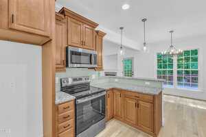 A modern kitchen with light wood cabinets, granite countertops, stainless steel stove and microwave, and pendant lights; open layout leads to a bright living area with large windows and wood flooring.