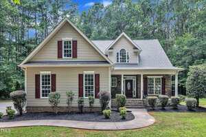 A beige two-story house at 2103 Troys Trail in Franklinton features dark red shutters, a covered front porch, and gabled roof, nestled among trimmed bushes and trees on a lawn bordered by a curved driveway.