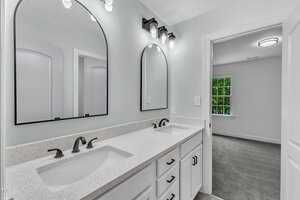 A modern bathroom with a double-sink vanity, white countertop, black fixtures, two arched mirrors, and overhead lighting. An open door leads to a carpeted room with a window showing greenery outside.