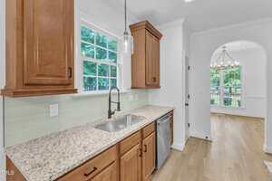 A modern kitchen features wooden cabinets, a granite countertop, a stainless steel sink with a pull-down faucet, a dishwasher, light green subway tile backsplash, and a window overlooking trees. An adjacent dining area is visible.