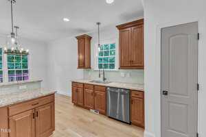 A modern kitchen with light wood flooring, brown wooden cabinets, granite countertops, stainless steel dishwasher, double windows above the sink, and a closed gray door on the right. A dining area is visible in the background.