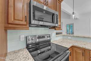 A modern kitchen features a stainless steel electric stove with a glass cooktop and an over-the-range microwave. The countertop is granite, and the backsplash is a light glass tile. Wooden cabinets are above and beside the appliances.