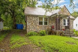 A small stone house with a front porch, iron railing, and a single red chair. The yard is grassy with some plants near the house, and a dirt driveway runs along the left side with a blue tarp visible in the backyard.