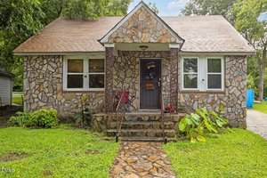 A small stone house at 630 E Avenue in Henderson with a front porch, stone steps, and a wooden door. Two chairs sit on the porch, flanked by plants along the steps, and a green lawn surrounds the home.