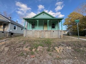 A small green house at 402 Spring Court in Henderson sits on a raised concrete foundation with a front porch. The sloped, patchy yard fronts a street sign reading "Spring St." with neighboring homes visible on both sides.