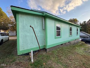A small, light green house with darker green trim, raised on a concrete foundation, sits on a grassy lot. There are several cars parked nearby and a rusty metal pipe extends from the ground in front of the house.