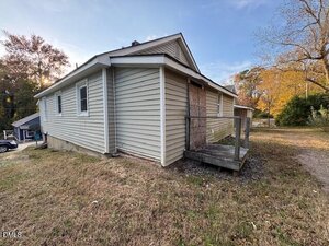 A small, single-story house with light-colored siding and a boarded-up door sits at 410 Spring Court in Henderson. It features a small wooden porch and is surrounded by a patchy, grassy yard with trees in the background.