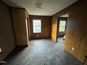A small, empty room with wood-paneled walls, two windows, a white ceiling light fixture, and a visibly worn, dirty floor. The space is dimly lit by natural light from the windows.