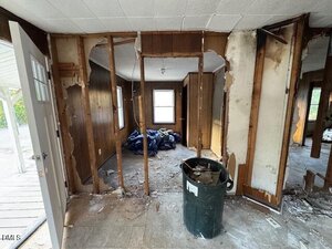 A room with exposed wall studs and damaged walls, debris on the floor, a trash bin filled with rubble, and blue bags in the adjoining room. The area appears to be under renovation or in disrepair.