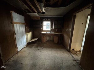 A dimly lit, empty kitchen with damaged flooring, exposed ceiling beams, missing cabinet doors, and visible debris. A single window is above the sink, and a white door is on the left wall.