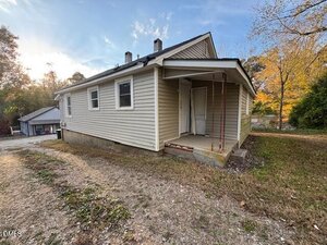 A small, single-story house with light-colored siding and a sloped roof. There is a covered entryway with metal posts and a concrete step. The yard is mostly dirt and grass with trees in the background.