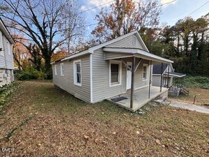 A small, single-story house with beige siding, a front porch with a white door, and a sloped roof. The house is surrounded by grass and trees with autumn leaves.