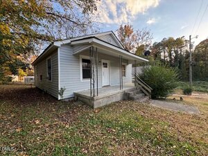 A small, single-story white house with a front porch, black metal posts, concrete steps, and a wooden railing. The yard has patchy grass, scattered leaves, and is surrounded by trees.