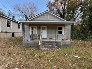 A small, single-story house with a covered front porch, two steps leading to a white door, and siding exterior. The yard has scattered leaves, and there are trees and another house in the background.