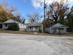 Three small single-story houses with front porches line a slightly sloped street on 521 Spring Court in Henderson. Leafless trees and autumn foliage fill the background, while power lines stretch overhead beneath a partly cloudy sky.