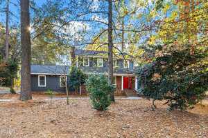A two-story gray house with white trim and a red front door is surrounded by tall trees and a yard covered with dry pine needles and fallen leaves on a sunny day.