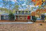 Charming two-story house at 109 Dogwood Circle, Butner, with dark blue-gray siding, red double front doors, brick steps and base. Surrounded by trees with autumn leaves and a yard of pine needles. Curved walkway leads to the entrance.
