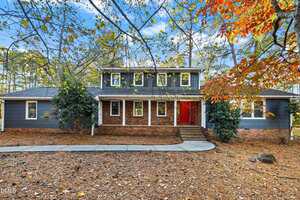 Charming two-story house at 109 Dogwood Circle, Butner, with dark blue-gray siding, red double front doors, brick steps and base. Surrounded by trees with autumn leaves and a yard of pine needles. Curved walkway leads to the entrance.