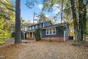 A two-story house with dark gray siding, white trim, and a brick foundation is surrounded by tall trees and fallen pine needles. The home has multiple windows and a covered front porch with columns.