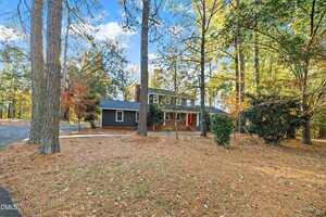 A two-story dark gray house with a red front door is surrounded by tall pine trees and a large, wooded yard covered in pine needles. The driveway curves past the house, which sits in a forested area under a blue sky.