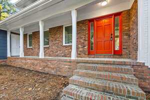 A brick house with a covered front porch, white columns, several windows, and a red front door with glass panels. Brick steps lead up to the entrance. The house is surrounded by mulch and natural landscaping.