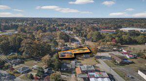 Aerial view of a semi-rural area with houses, trees, and roads. One property in the center is highlighted with an orange outline, indicating a specific lot or building within the neighborhood.