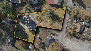 Aerial view of two adjacent, L-shaped empty lots outlined in orange at 210/212 N Shank Street, Henderson. The lots are cleared with grass and bare ground, surrounded by trees, a few houses, parked vehicles, and a dirt road.