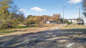A dirt road runs through a grassy, open lot with scattered trees. Utility poles and wires are visible. Houses and sheds are seen in the background under a partly cloudy sky. Pieces of wood lay on the ground in the foreground.