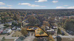 Aerial view of a suburban neighborhood with a marked plot of land outlined in orange in the center; surrounding area features houses, trees, and a road running along the left edge of the image.