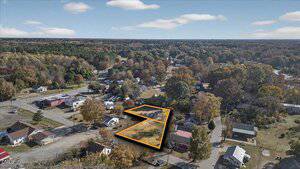 Aerial view of a residential neighborhood with tree-lined streets. Two adjacent empty lots are highlighted in orange near the center of the image, surrounded by houses and roads. Dense forest is visible in the distance.