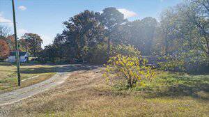 A gravel driveway curves through a grassy area with sparse vegetation. Leafy trees border the scene, and a few houses are visible in the background under a clear blue sky.
