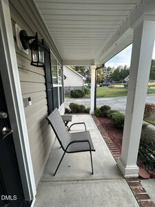 A front porch with two metal chairs and a small side table sits under a white roof. The porch is bordered by shrubs and overlooks a driveway and residential street.