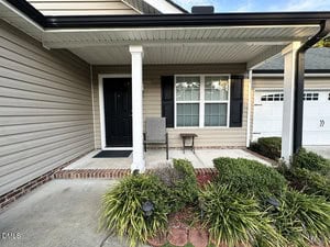 A covered front porch with a black door, beige siding, two windows with black shutters, a gray chair, a small table, and landscaped greenery in front of the porch.